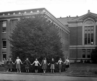 Coeds encircling the giant holly tree behind what is now Kidder Hall, December 1940. The women pictured were members of the Theta Sigma Phi national women's honor society in journalism.